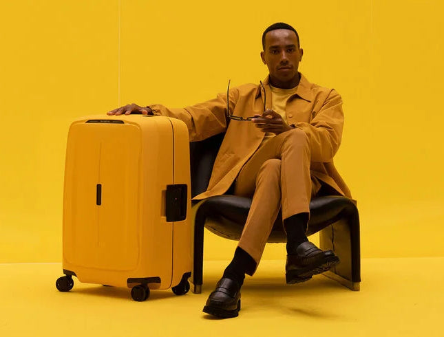 en mann i oker farget antrekk sitter i en stol og lener hånden på en gul samsonite essens koffert i hardplast med klipslås