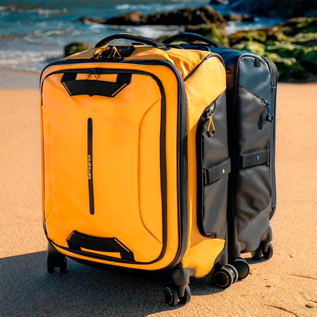 bilde av to trillekofferter i vannavvisende materiale på en strand en solfylt dag, ene kofferten er gul og den andre blå. 