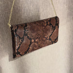 Abro Python clutch skulderveske i mørk sjokoladebrunfarget skinn med forgyllet kjettingrem. Vesken vises frem i kort video foran en grå/beige bakgrunn. 