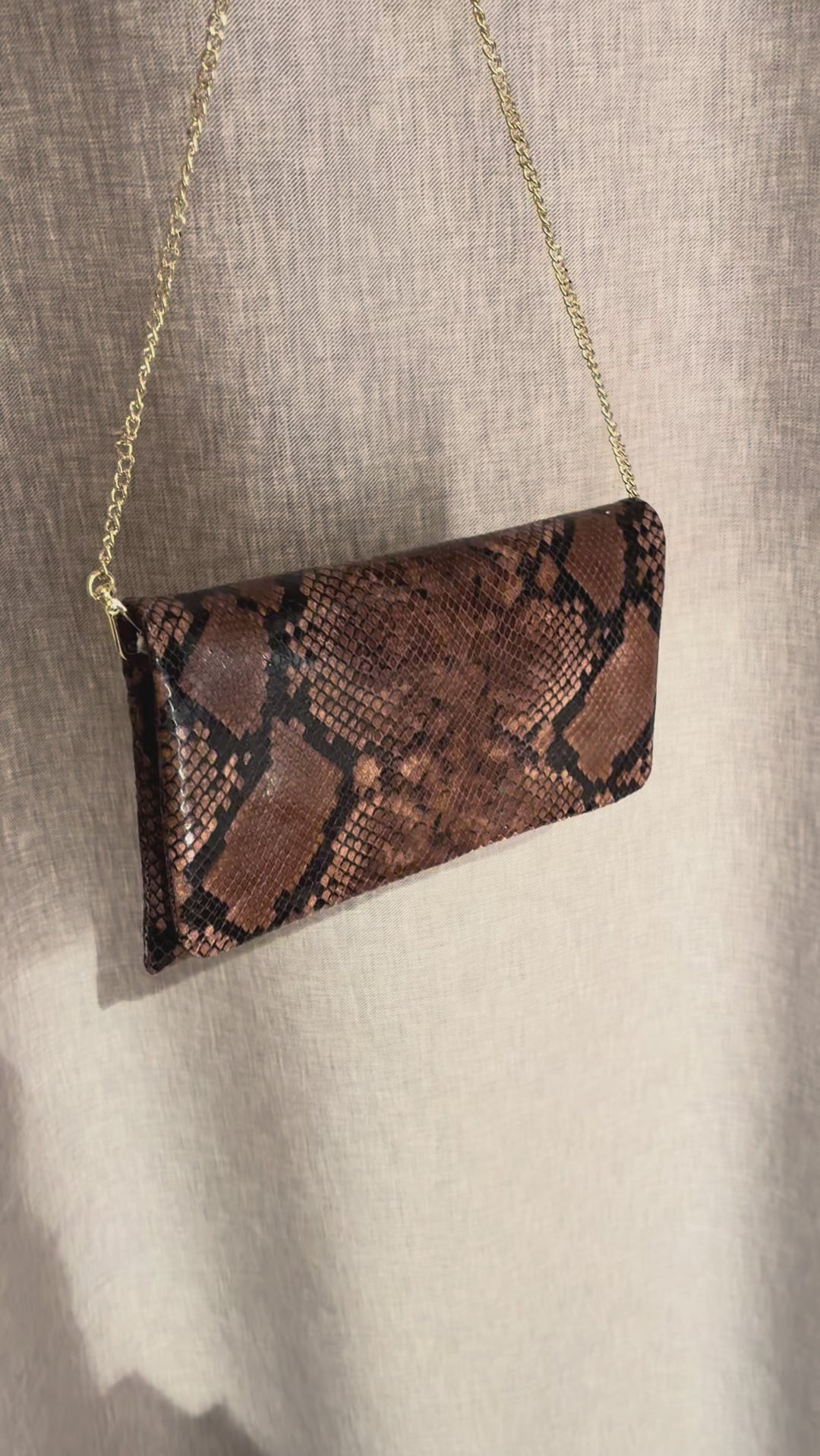 Abro Python clutch skulderveske i mørk sjokoladebrunfarget skinn med forgyllet kjettingrem. Vesken vises frem i kort video foran en grå/beige bakgrunn. 