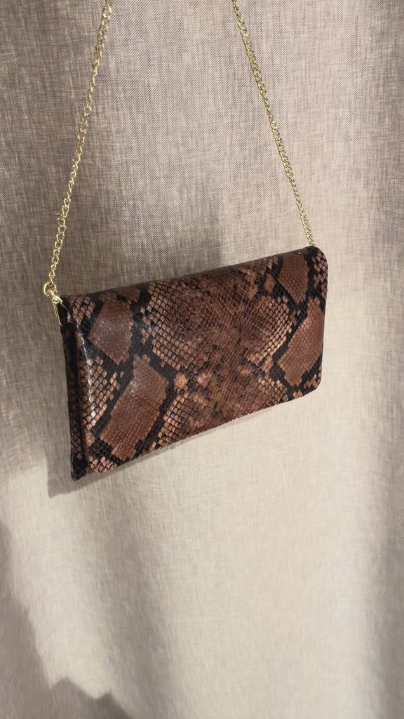Abro Python clutch skulderveske i mørk sjokoladebrunfarget skinn med forgyllet kjettingrem. Vesken vises frem i kort video foran en grå/beige bakgrunn. 