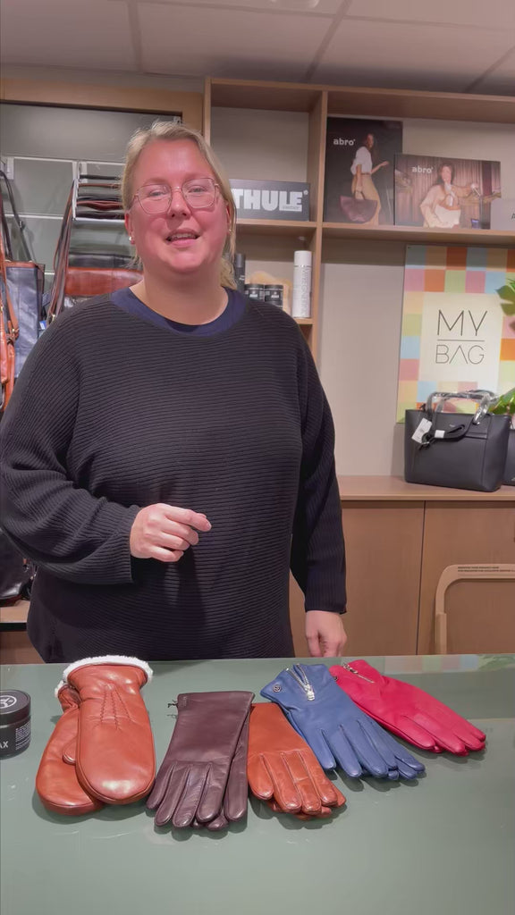 video med en dame i butikk som viser hvordan en skinnhanske som passer perfekt skal sitte på hånden. Hun har forskjellige skinnhansker i forskjellige farger liggende på bordet foran seg. Hun demonstrerer passformen med noen blå skinnhansker fra Adax