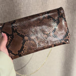 Abro Python clutch skulderveske i mørk sjokoladebrunfarget skinn med forgyllet kjettingrem. Vesken vises frem i kort video hvor vesken holdes i en hånd og vinkles i lyset for å vise skinnets glansfulle egenskaper