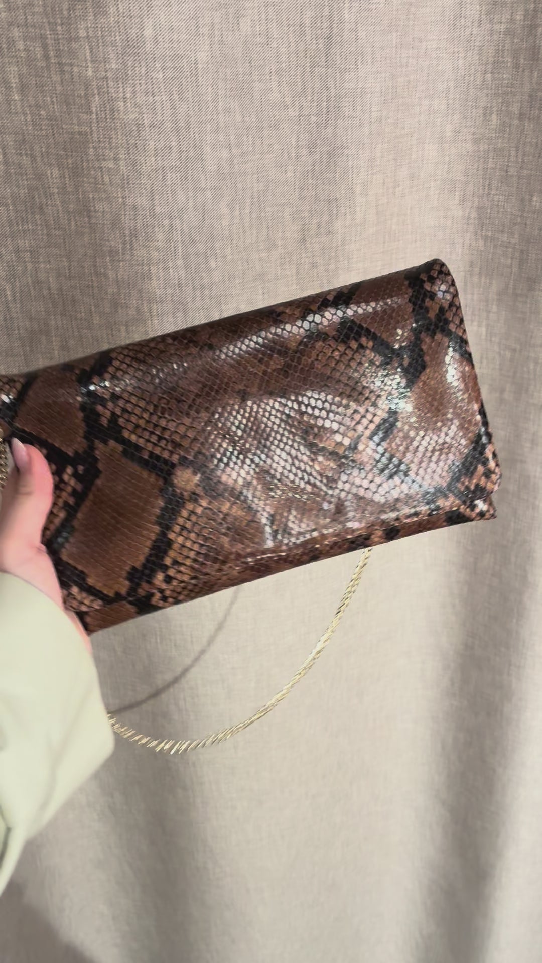 Abro Python clutch skulderveske i mørk sjokoladebrunfarget skinn med forgyllet kjettingrem. Vesken vises frem i kort video hvor vesken holdes i en hånd og vinkles i lyset for å vise skinnets glansfulle egenskaper