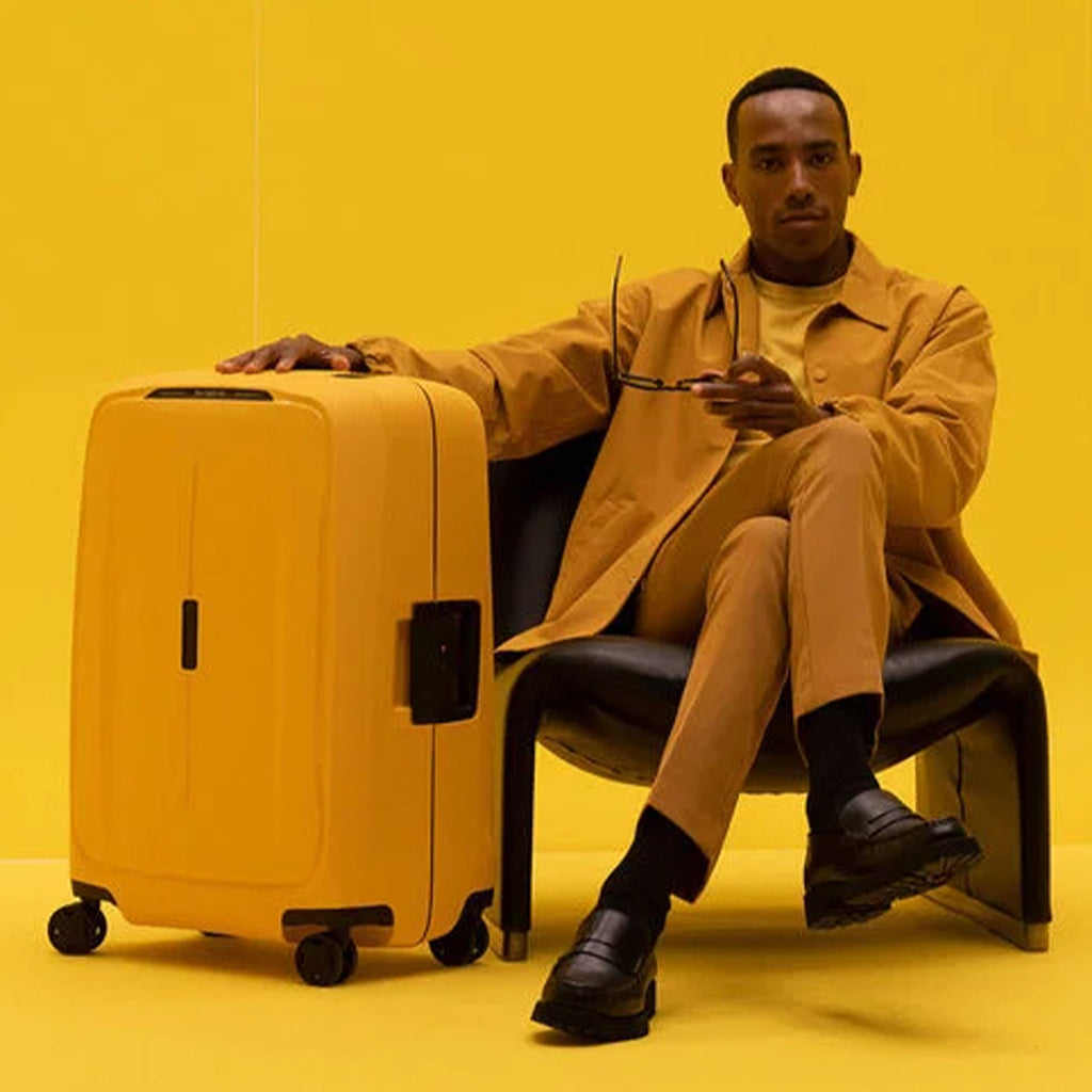 bilde av en mann i okerfarget dress, sittende på en skinnstol med gul bakgrunn, vedsiden står det en gul samsonite koffert