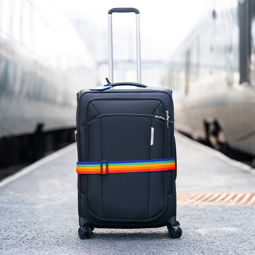 bilde av en sort koffert med en regnbuet koffertstropp på langs, stående mellom to tog på en togstasjon
