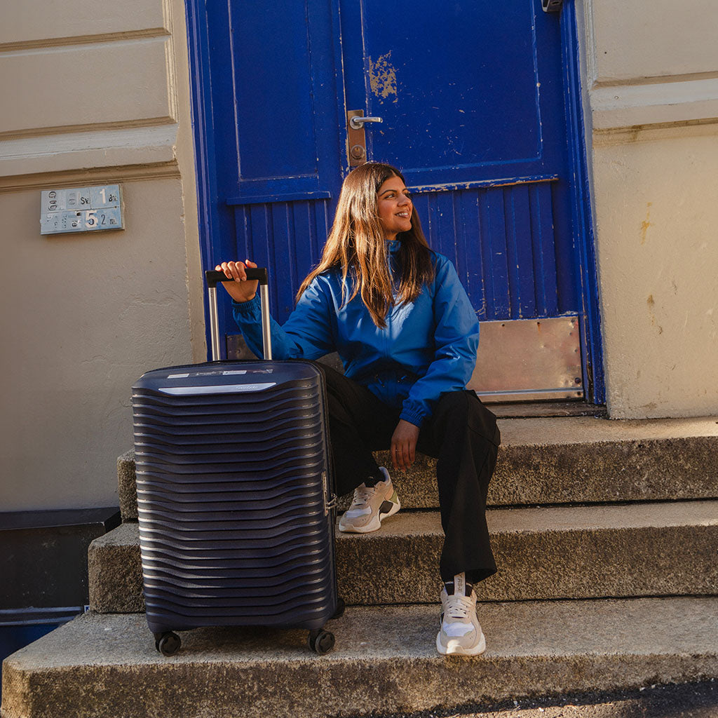 bilde av en jente sittende på en trappe utenfor en blå dør, smilende i solen mens hun har en blå koffert stående på siden